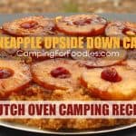 This is a photo of a Dutch Oven Pineapple Upside Down Cake Camping Recipe by CampingForFoodies. The moist cake is covered with gooey pineapples dotted with cherries sitting on a table in the mountains at a remote camping site.
