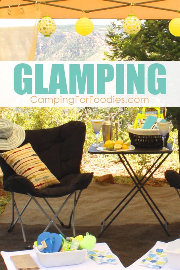 Give Yourself A Pedi On Your DIY Glamping Trip by CampingForFoodies is an image of a comfy camp chair and a bucket filled with accessories to do a DIY pedicure, setting on a rug under a camping canopy that is set up in the area of a forest with a beautiful rock wall in the backdrop. Alongside the chair is a folding camping table holding a pretty basket filled with magazines setting next to a cocktail shaker and a plastic martini glass filled with freshly shaken lemonade.