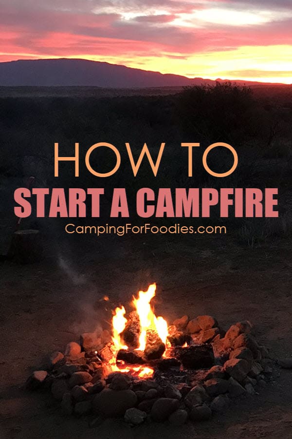 How to start a camp fire by CampingForFoodies features a campfire burning at twilight with a beautiful sunset with a pink, orange and yellow sky filled with fluffy clouds and text over the image that reads how to start a campfire.