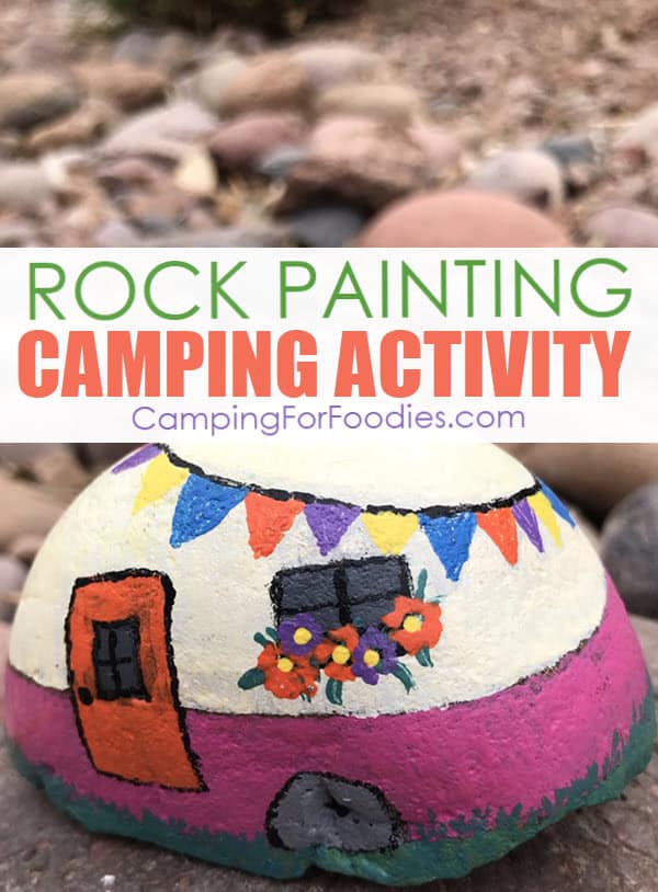 Rock Painting Camping Activity by CampingForFoodies features a brightly colored painted rock craft project that looks like a little cream and pink retro camper decorated with blue, orange, purple and yellow flags and orange and purple flowers in a window box under the trailer’s window the orange door welcomes visitors and round river rocks are piled in the background with text over the image that reads rock painting camping activity.