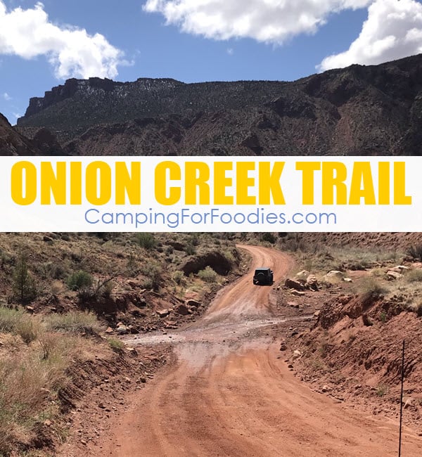 Onion Creek Trail Moab Utah by CampingForFoodies features a jeep driving uphill on the curving Onion Creek Road motorized vehicle trail after passing through the muddy creek crossing with gorgeous red rock landscape and mountains in the distance under a sunny blue sky with white fluffy clouds and text over the image that reads Onion Creek Trail.