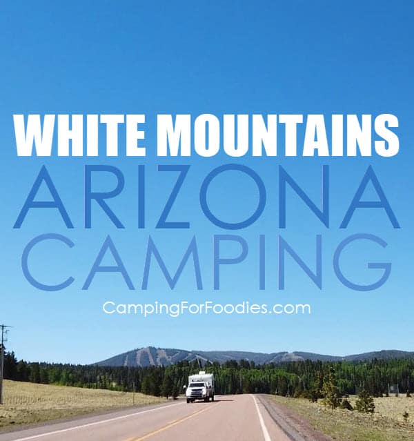 Camping In Arizona White Mountains by CampingForFoodies features a truck pulling a camper down a scenic road in the summer in Arizona with evergreen trees and mountains with ski slope runs under a bright blue sky in the background with text over the image that reads White Mountains Arizona Camping.