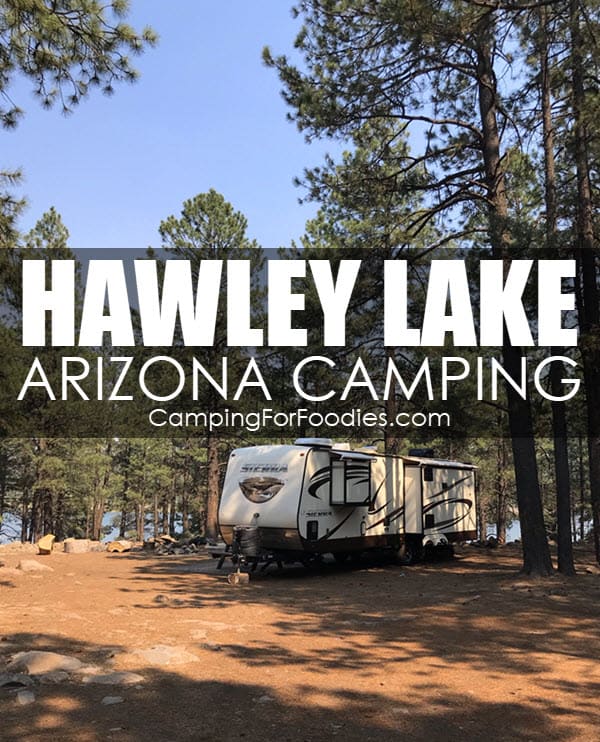 Hawley Lake Arizona Camping by CampingForFoodies features a travel trailer parked at a wooded campsite under a bright blue sky with a scenic lake in the background with text over the image that reads Hawley Lake Arizona Camping.