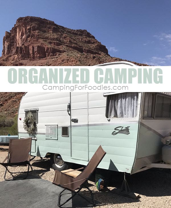 Organized Camping With Camping Organization Ideas For How To Stay Clean And Organized Throughout Your Entire Trip by CampingForFoodies features a retro Shasta camper with a white and light green color scheme at two camp chairs neatly arranged on an outdoor carpet in front of the door with a cloth-covered picnic table in the background under a blue sky and scenic red rocks in the background at a beautiful campsite with text over the image that reads organized camping.