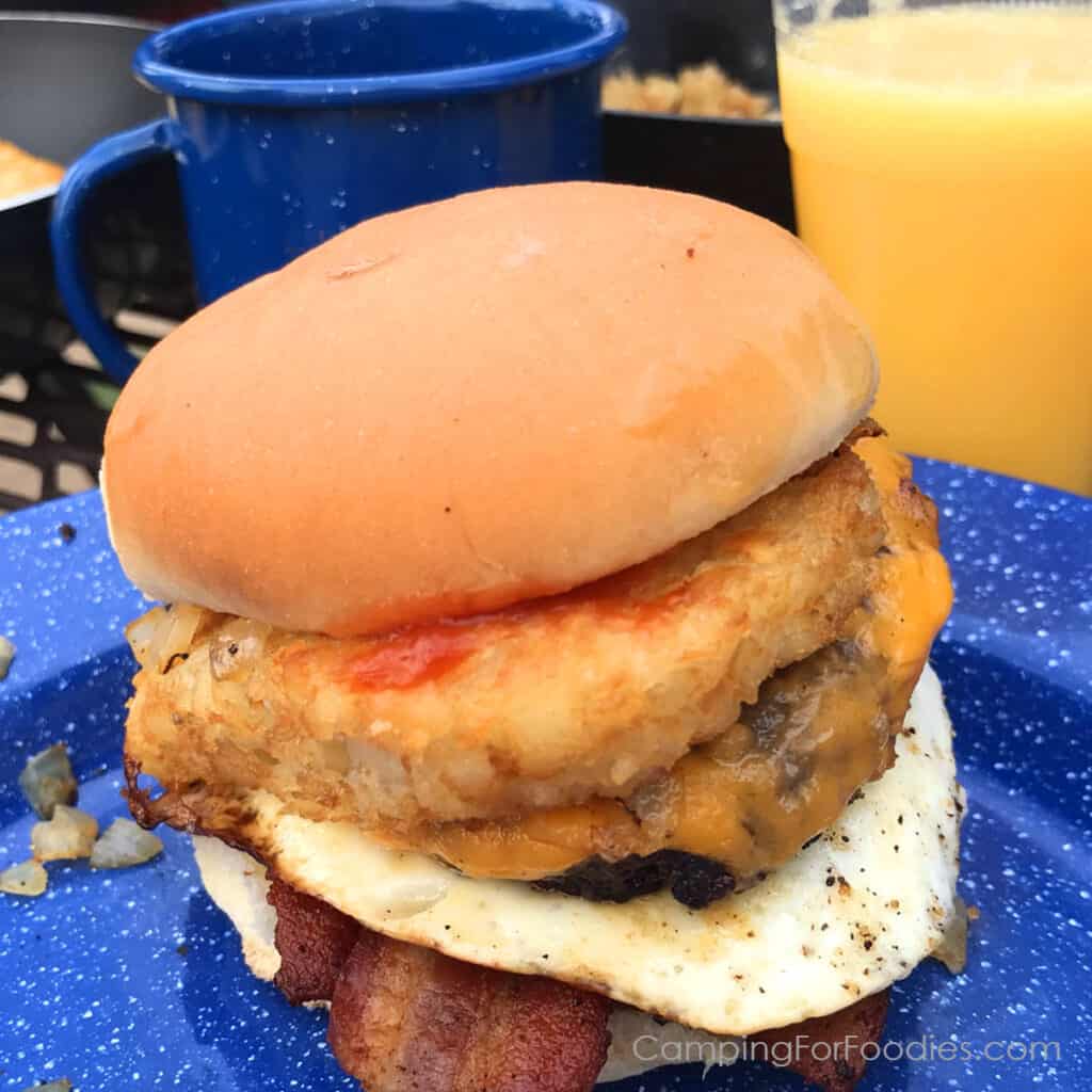 All Day Breakfast Burger Recipe by CampingForFoodies features a breakfast sandwich on a blue camping plate. The potato hamburger bun holds the layers of ingredients including maple bacon, a fried egg, sausage patty with melted cheddar cheese, a golden brown hash brown patty, sauteed onions and ketchup. In the background is a cup of coffee and glass of fresh orange juice.