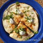 Camping Breakfast Quesadillas by CampingForFoodies features a golden brown, toasted breakfast quesadilla on a blue camping plate ready to be served at the campsite. The filling includes eggs, chorizo sausage and cheese with toppings of sour cream and fresh green cilantro.