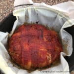 Dutch Oven Meatloaf by CampingForFoodies features a bacon and glazed-topped meat loaf nestled in a Dutch oven parchment paper liner that is set inside a cast iron camp Dutch oven ready to be sliced and served at the campsite.