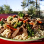 Beef Broccoli And Carrot Camping Stir Fry by CampingForFoodies features a plate full of food sitting on a picnic table at a campsite, ready to be served for dinner. The beef, broccoli and carrot stir fry is sitting on a bed of brown rice and topped with sesame seeds. In the background is a beautiful forest under a pretty blue sky on a sunny day.
