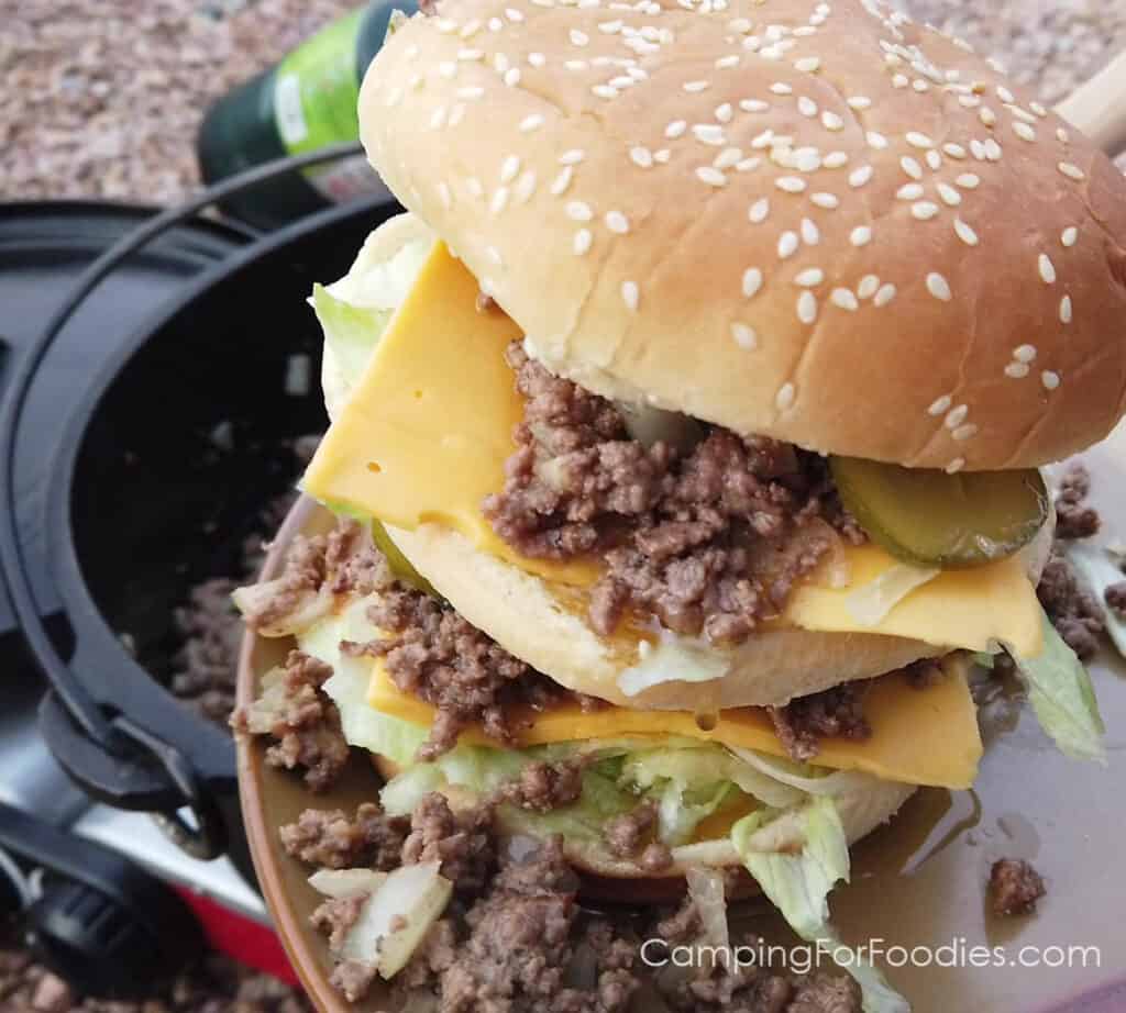 Big Mac Sloppy Joes by CampingForFoodies features a double decker sandwich on a sesame seeded bun filled with sloppy joe meat and a copycat version of McDonald’s secret sauce, American cheese, pickles and shredded lettuce. The meal is ready to be served for dinner at the campsite. A cast iron Dutch oven and propane fuel canister on a camp stove are in the background.