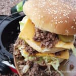 Big Mac Sloppy Joes by CampingForFoodies features a double decker sandwich on a sesame seeded bun filled with sloppy joe meat and a copycat version of McDonald’s secret sauce, American cheese, pickles and shredded lettuce. The meal is ready to be served for dinner at the campsite. A cast iron Dutch oven and propane fuel canister on a camp stove are in the background.