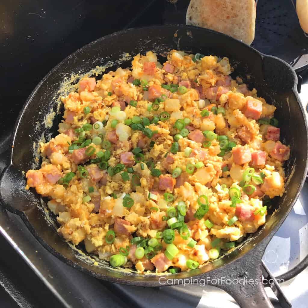 Camping Breakfast Skillet by CampingForFoodies features a cast iron skillet on a camp stove filled with a one pot meal of ham, eggs, potatoes, cheese and green onions ready to be served at the campsite, in the background are English muffins being toasted on a camp stovetop toaster.