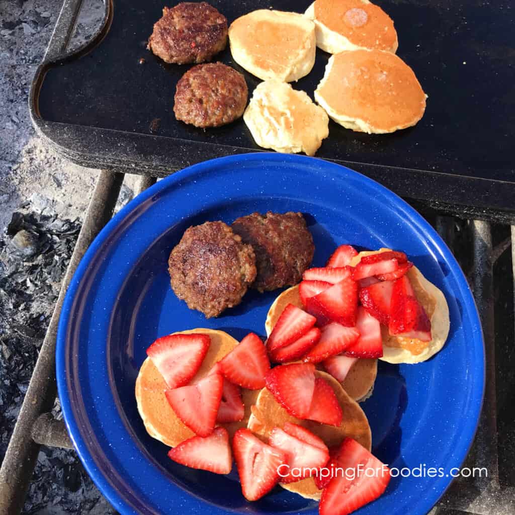 Easy Camping Pancakes by CampingForFoodies features a blue camping plate filled with golden brown pancakes topped with bright red strawberries served on the side with spicy breakfast sausage patties, the plate is sitting on a campfire grate, in the background on the same grate is a black cast iron griddle filled with more pancakes and sausage patties.