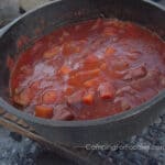 Campfire Stew In Rich Tomato Sauce by CampingForFoodies features a Dutch oven over a campfire filled with a tender beef stew in a rich tomato based sauce with carrots and potatoes ready to be served at the campsite.