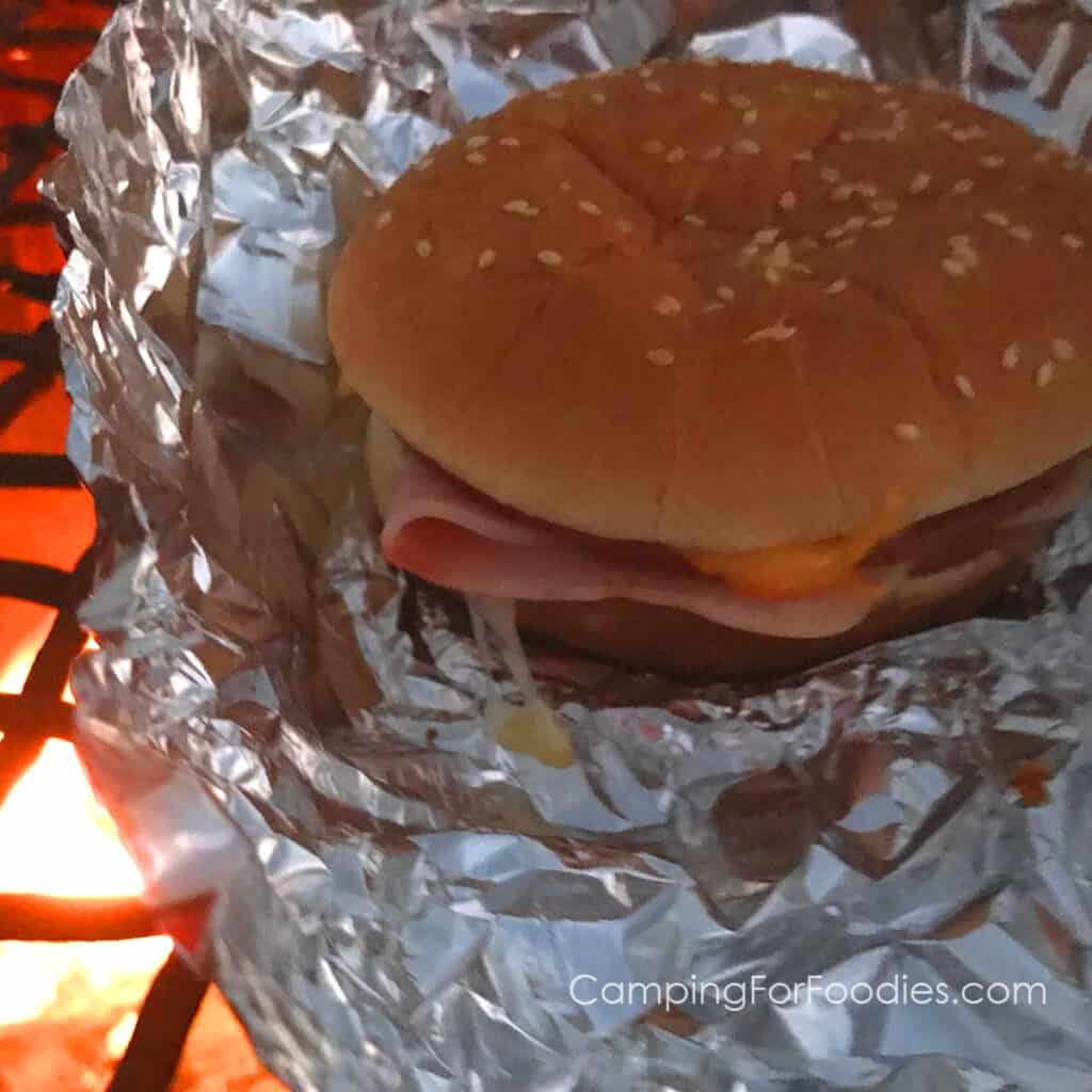 Chuckwagon Sandwich Recipe by CampingForFoodies features a sesame seeded hamburger bun filled with salami, bologna, ham, American and Swiss cheese slices set on a piece of aluminum foil on top of a campfire cooking grate over a red, orange and yellow-flamed campfire.