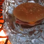 Chuckwagon Sandwich Recipe by CampingForFoodies features a sesame seeded hamburger bun filled with salami, bologna, ham, American and Swiss cheese slices set on a piece of aluminum foil on top of a campfire cooking grate over a red, orange and yellow-flamed campfire.