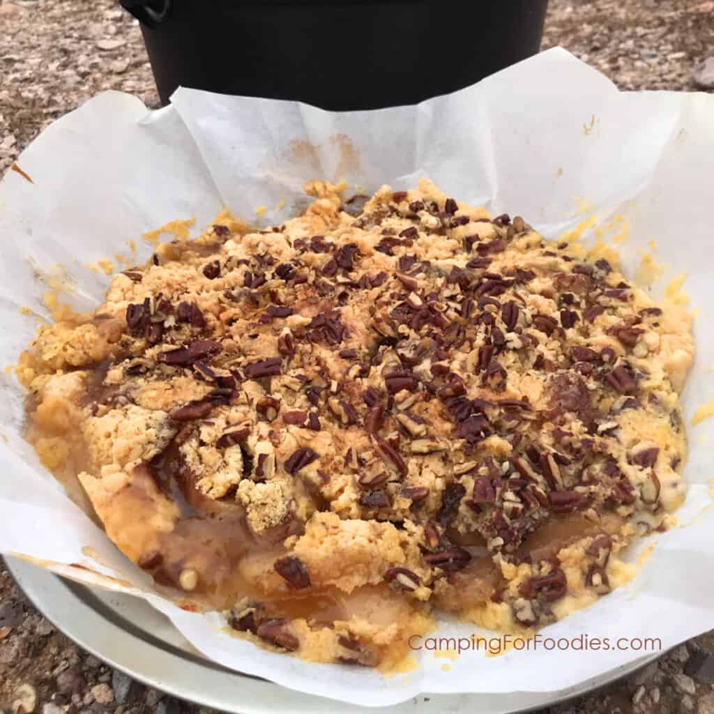 Dutch Oven Apple Dump Cake by CampingForFoodies features a golden brown dessert set in a parchment paper liner with apple pie filling that is sprinkled with a boxed cake mix, butter and pecans then baked till bubbly and golden brown in a camp Dutch oven which is sitting the background. The dessert is ready to be served at the campsite.