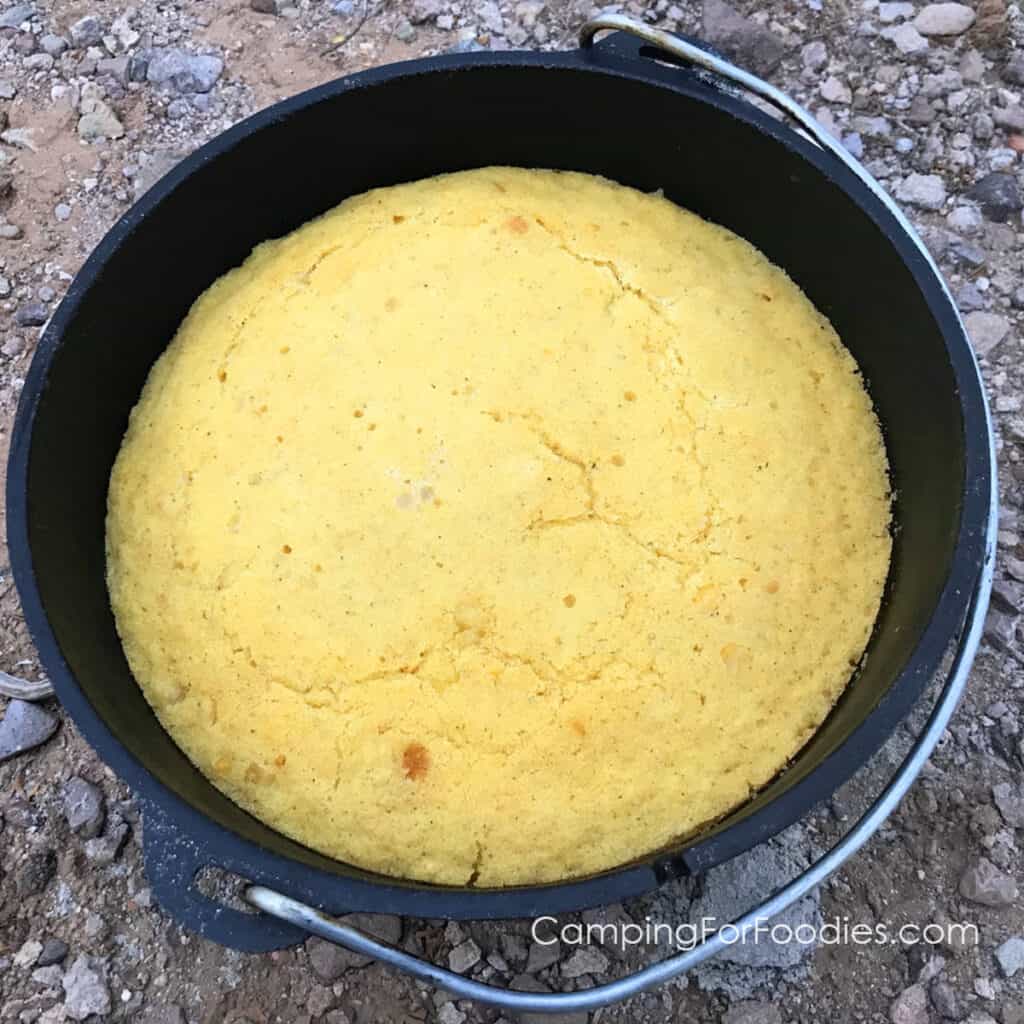 Dutch Oven Cornbread by CampingForFoodies features a black cast iron Dutch oven filled with a freshly baked recipe of golden-brown cornbread, set on the ground to cool, waiting to be served at the campsite.