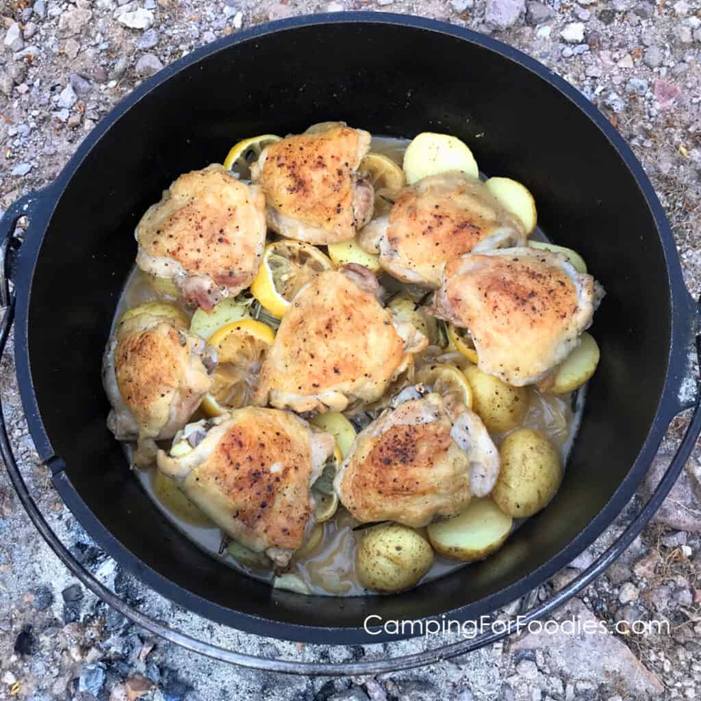 Dutch Oven Lemon Chicken by CampingForFoodies features a black cast iron Dutch oven filled with golden brown, crispy chicken thighs on top of a bed of tender petite potatoes, lemon slices and fresh rosemary sprigs in a smooth pan sauce. The pot is sitting on the ground, dinner is ready to be served at the campsite.