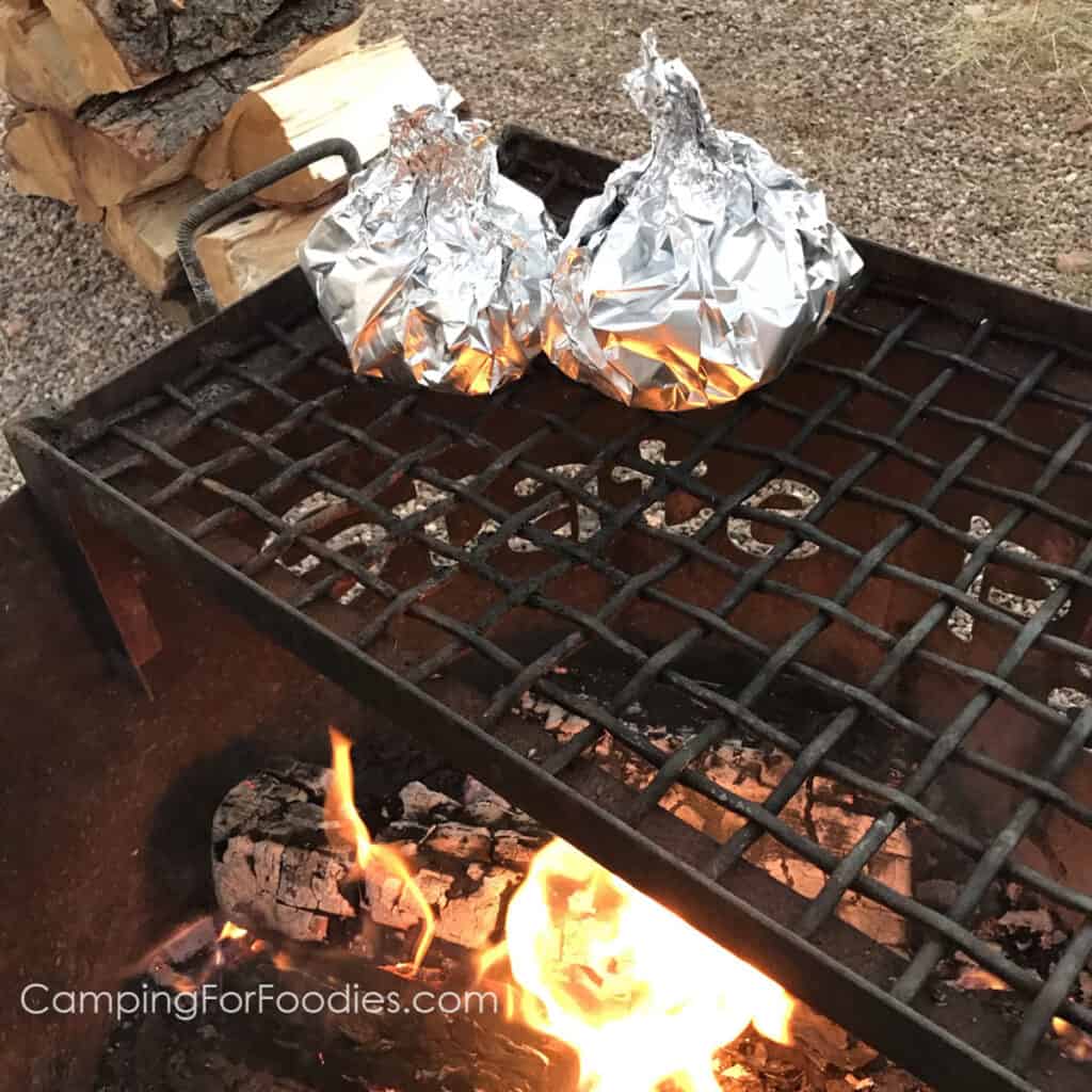 Foil Wrapped Chuck Wagon Sandwich Recipe by CampingForFoodies features two foil packets set on top of a campfire cooking grate over a orange and yellow-flamed campfire and a pile of split firewood in the background.