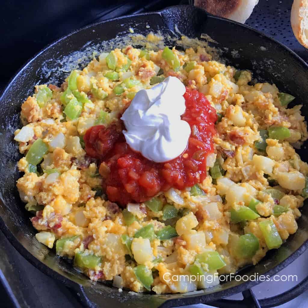 Hungry Man Breakfast by CampingForFoodies features a black cast iron skillet on a propane camp stove filled with a hearty breakfast ready to be served. The one pot meal includes bacon, southern style hash browns, green bell pepper, onion, cheddar cheese, scrambled eggs and is topped with salsa and sour cream. In the background is a camp toaster with golden brown English muffins and a slice of toasted wheat bread.