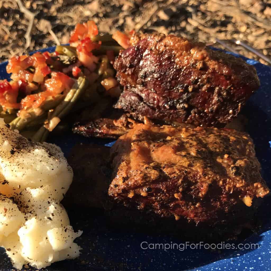 Pellet Smoked Short Ribs by CampingForFoodies features a blue camping plate filled with food ready to be served at the campsite. The English-style cut short ribs have a golden brown color after being smoked in a pellet grill. Next to the ribs are smoked green beans with bacon and fluffy mashed potatoes sprinkled with freshly ground black pepper.