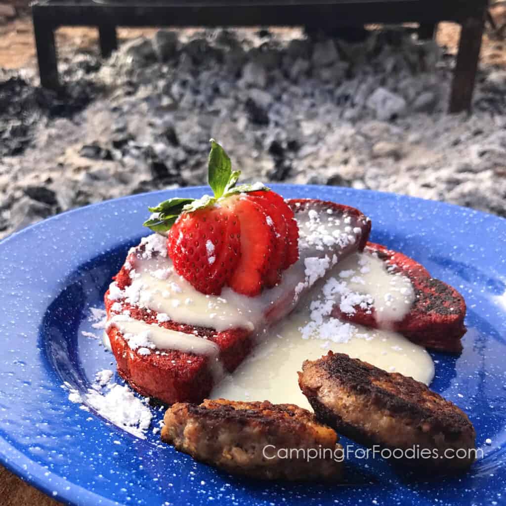 Red Velvet French Toast by CampingForFoodies features a blue camping plate filled with two slices of red velvet French toast topped with sweet cream cheese glaze, powdered sugar and a sliced and fanned strawberry next to two golden brown breakfast sausage patties and a campfire grate set inside a campfire ring in the background.