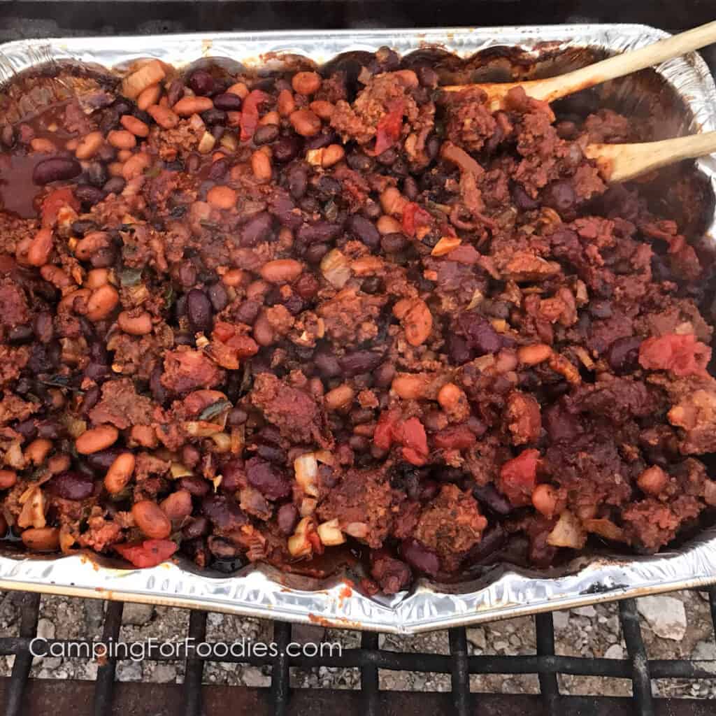 Smoked Ground Beef Chili by CampingForFoodies features an aluminum foil pan filled with smoked chili that has been smoked in a portable pellet grill and includes hamburger, bacon, onion, garlic, jalapeno peppers, spices, three types of beans, tomatoes and sauce that is ready to be served at the campsite.