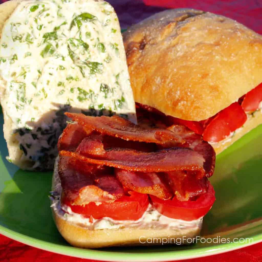 Bacon Basil Tomato Ciabatta Sandwich Recipe by CampingForFoodies features two ciabatta sandwiches on a green plate. One sandwich is closed next to another one that has the upper bun setting up against the lower bun that is piled with golden brown bacon and deep red tomato slices. Green and white basil mayonnaise is spread on the inside of the top and bottom buns.