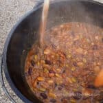 Dutch Oven Baked Beans Cast Iron 5-Bean Recipe by CampingForFoodies features a black cast iron camp Dutch oven filled with five types of beans in a homemade sauce being stirred with wooden spoons as steam rises from the pot. This baked beans with hamburger and bacon recipe is ready to be served at the campsite.