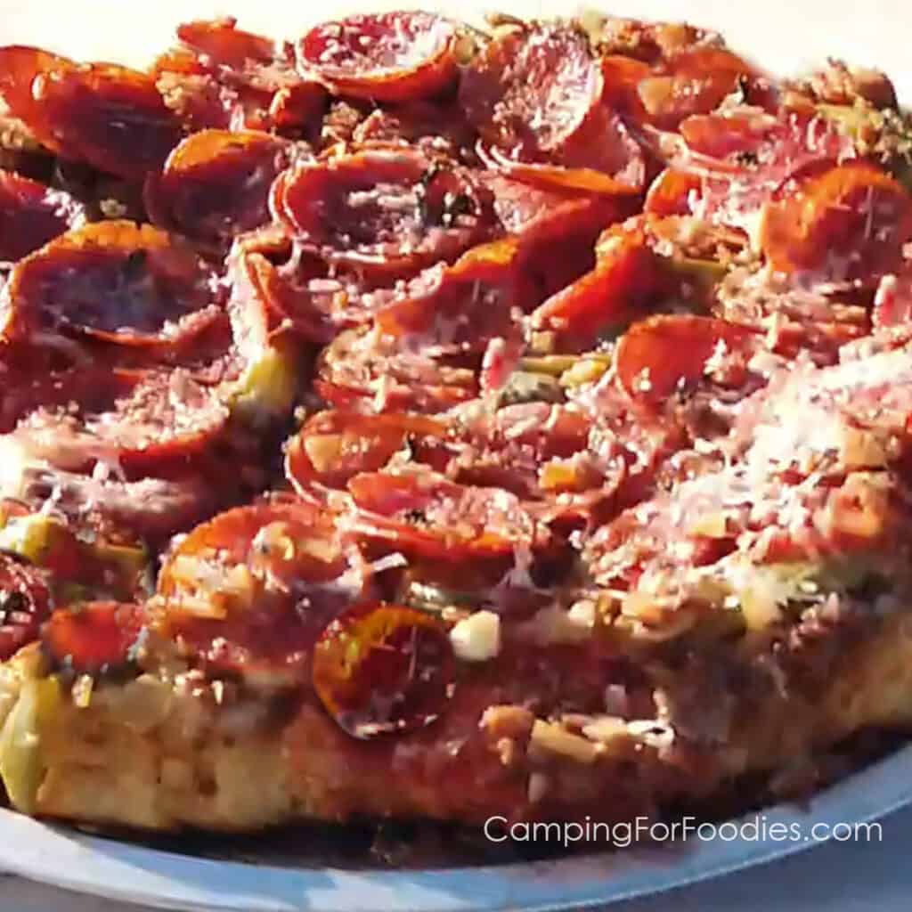 Dutch Oven Upside Down Pizza by CampingForFoodies features a fun twist on a classic deep dish pan style pizza that is made in a cast iron camp Dutch oven and then inverted onto a pizza serving tray. The pizza has bright red pepperoni, fresh basil, shredded mozzarella and parmesan cheeses and is ready to be served at the campsite.