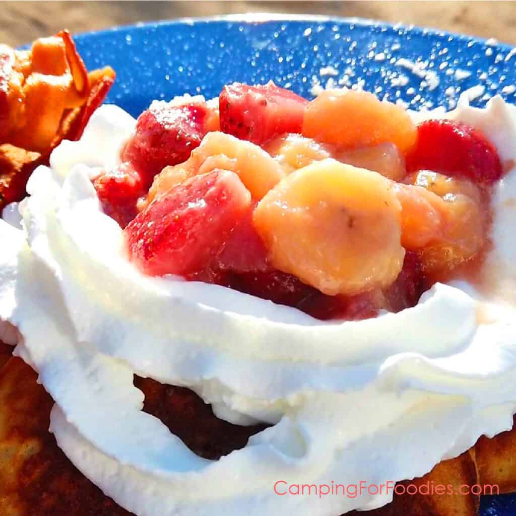 Strawberry Banana Compote by CampingForFoodies features a blue camping plate with strawberry banana compote and fluffy whipped cream topping a stack of pancakes. The red strawberries and golden banana slices glisten with the sweet syrup under a sunny sky as the plate of food is ready to be served at the campsite.