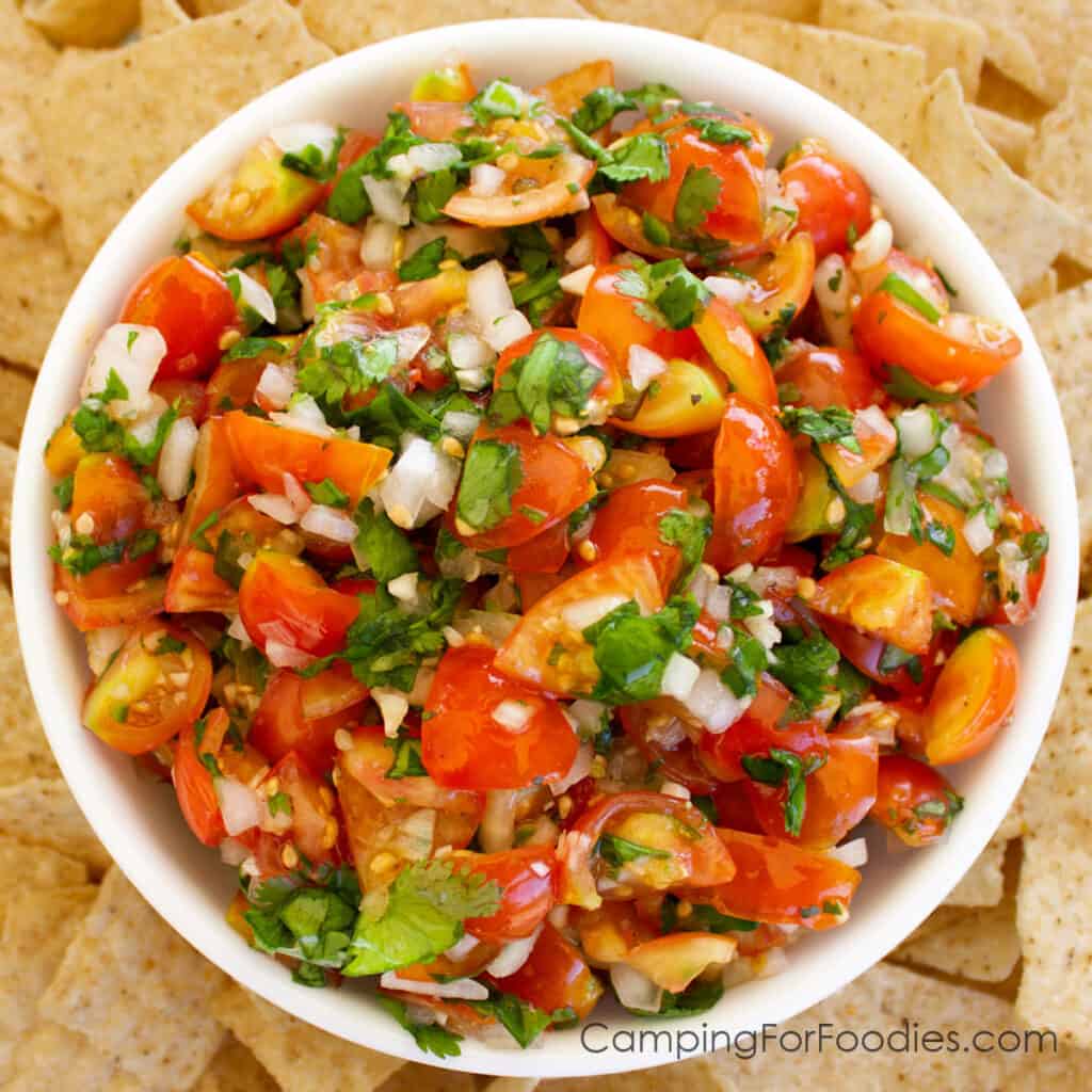 Sweet Onion Salsa Recipe by CampingForFoodies is a bowl full of red tomatoes, green cilantro and sweet white onions in a white serving bowl surrounded by a pile of tortilla chips ready to be served at the campsite.