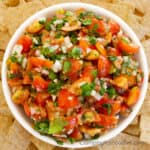 Sweet Onion Salsa Recipe by CampingForFoodies is a bowl full of red tomatoes, green cilantro and sweet white onions in a white serving bowl surrounded by a pile of tortilla chips ready to be served at the campsite.