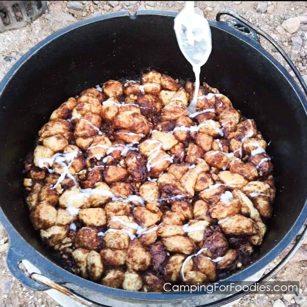 Dutch Oven Monkey Bread by CampingForFoodies features a black cast iron camp Dutch oven filled with hot-baked small pieces of refrigerator biscuits tossed in cinnamon, sugar and nuts being drizzled with a white powered sugar glaze.