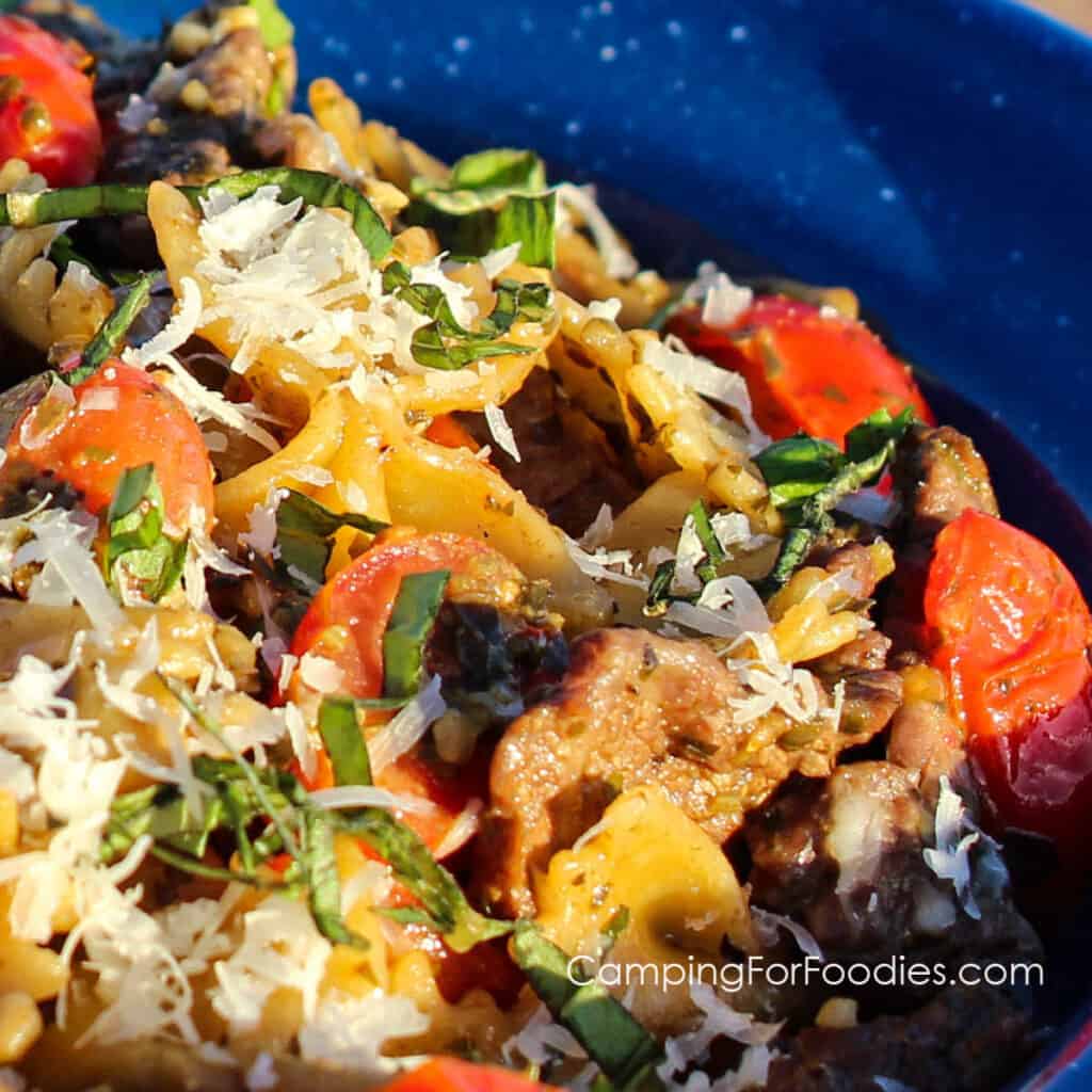 Pesto Steak Pasta Recipe by CampingForFoodies features a blue camping plate filled with a serving of pesto steak pasta that includes bow tie pasta, cherry tomatoes and strips of skirt steak tossed in pesto then topped with Parmesan cheese and fresh basil.