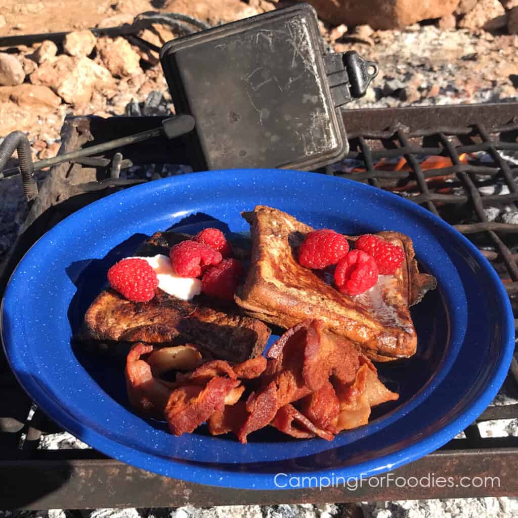 Pie Iron French Toast Recipe by CampingForFoodies features a blue camping plate filled with thick Texas-style French toast topped with melted butter, maple syrup and red raspberries with a campfire pie iron and flames in the campfire ring in the background.