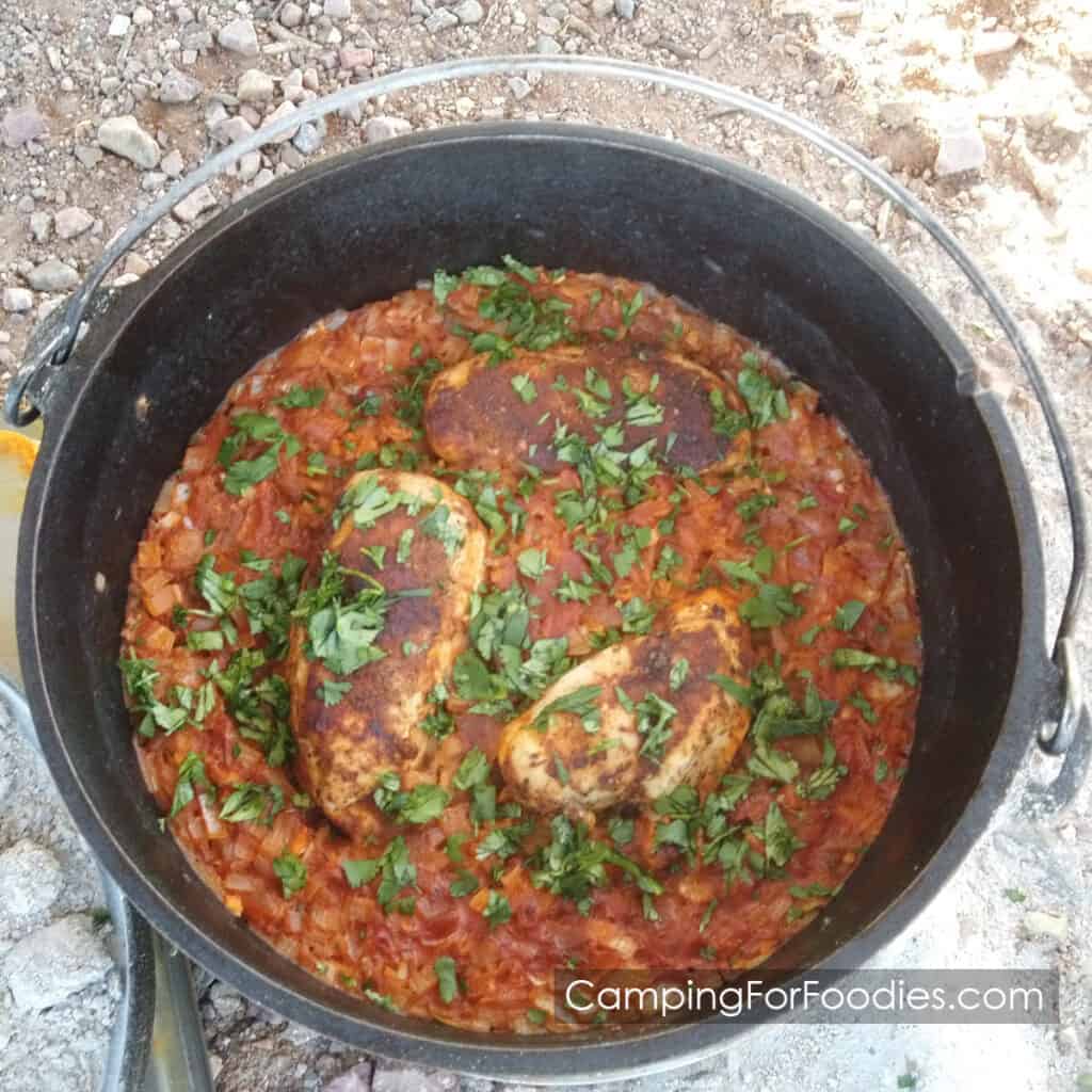 Dutch Oven Mexican Chicken Recipe by CampingForFoodies features a black cast iron Dutch oven filled with a freshly baked meal of browned chicken breasts and rice in a Mexican sauce topped with green cilantro.