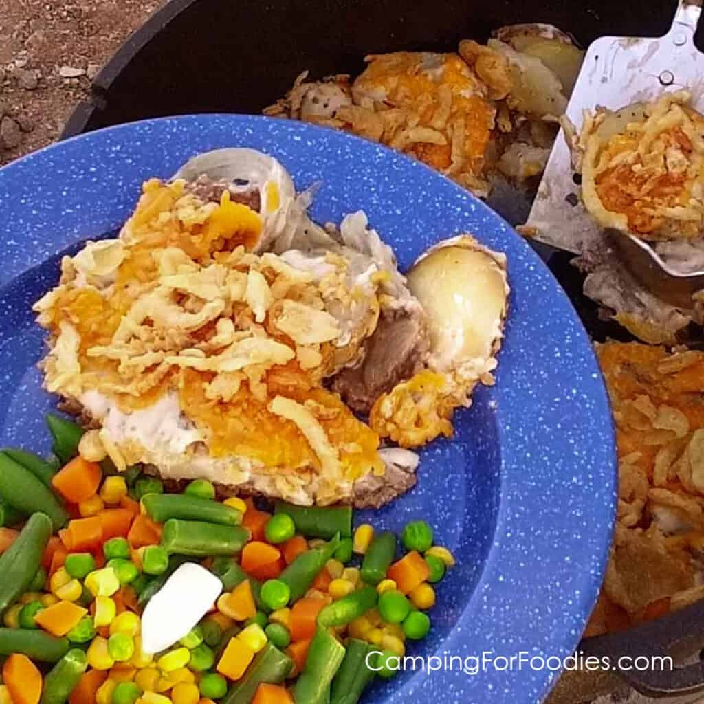 Hobo Casserole by CampingForFoodies features a blue camping plate filled with a serving of Hobo Casserole, ground beef patty topped with sliced potatoes, cream of mushroom soup, cheese and French fried onions, mixed veggies on the side and black cast iron Dutch oven in background with the remaining servings.