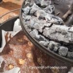 Campfire Brownies by CampingForFoodies features a black cast iron Dutch oven filled with freshly baked brownies dotted with mini Snickers candy bars as the lid filled with campfire coals is being removed from the top of the Dutch oven.