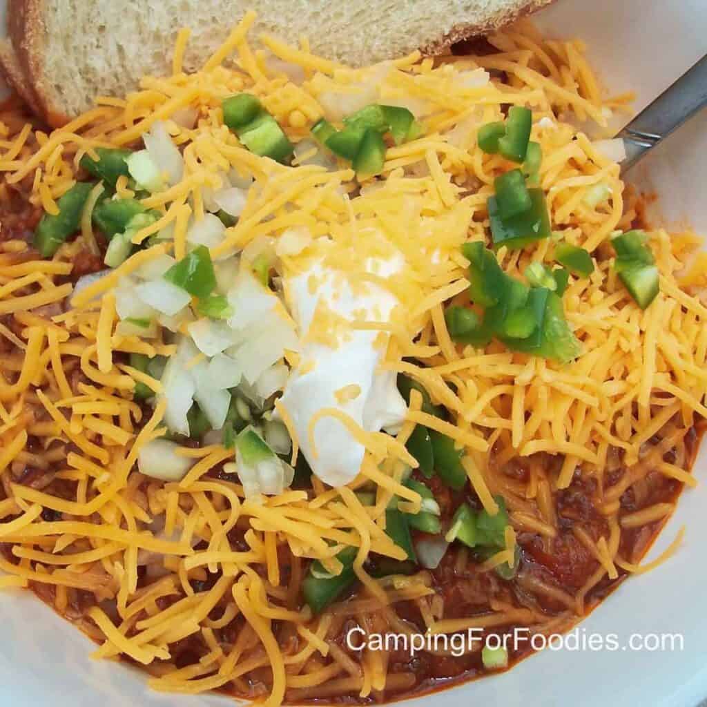 Campfire Chili by CampingForFoodies features a bowl of chili topped with cheddar cheese, sour cream, diced onions and jalapeno peppers with a spoon and slice of crusty bread ready to be served at the campsite.