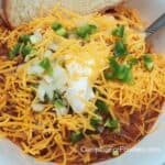 Campfire Chili by CampingForFoodies features a bowl of chili topped with cheddar cheese, sour cream, diced onions and jalapeno peppers with a spoon and slice of crusty bread ready to be served at the campsite.