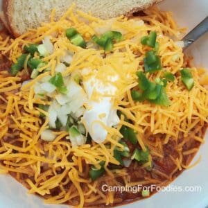 Campfire Chili by CampingForFoodies features a bowl of chili topped with cheddar cheese, sour cream, diced onions and jalapeno peppers with a spoon and slice of crusty bread ready to be served at the campsite.