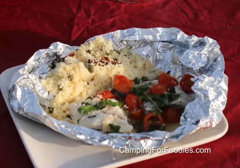 Campfire Fish Grilled In Foil Packets by CampingForFoodies features flaky white fish, red cherry tomatoes and ribbons of fresh green basil in an opened aluminum packet with a pile of couscous mixed with capers and sun-dried tomatoes ready for serving.