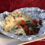 Campfire Fish Grilled In Foil Packets by CampingForFoodies features flaky white fish, red cherry tomatoes and ribbons of fresh green basil in an opened aluminum packet with a pile of couscous mixed with capers and sun-dried tomatoes ready for serving.