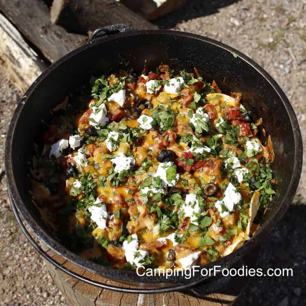 Campfire Nachos by CampingForFoodies features a black cast iron Dutch oven pot filled with nachos topped with melted cheese, fire roasted tomatoes, sour cream, black olives and cilantro with firewood logs in the background.