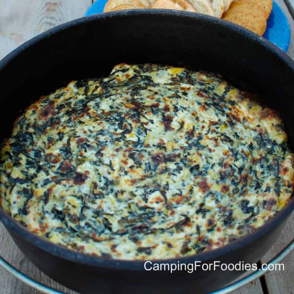 Campfire Spinach Dip by CampingForFoodies features a golden brown, hot cheesy spinach artichoke dip in a black cast iron Dutch oven on a wooden picnic table with crackers on a blue camping plate in the background. It's one of the best easy camping recipes that can be served during happy hour while the entire family is sharing the good times they had that day.