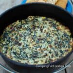 Campfire Spinach Dip by CampingForFoodies features a golden brown, hot cheesy spinach artichoke dip in a black cast iron Dutch oven on a wooden picnic table with crackers on a blue camping plate in the background.