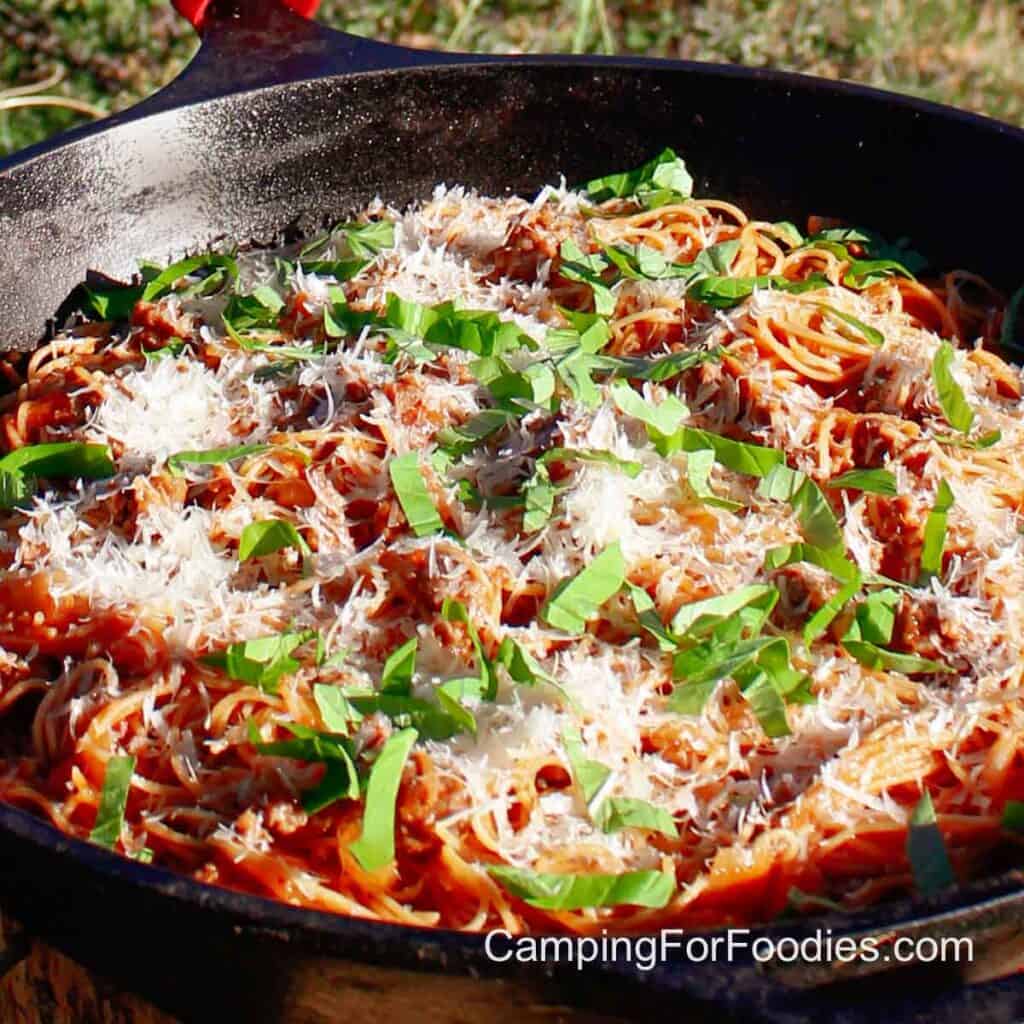 Camping Spaghetti by CampingForFoodies features a big cast iron skillet filled with pasta cooked in a sausage tomato sauce and topped with fresh basil and parmesan cheese.