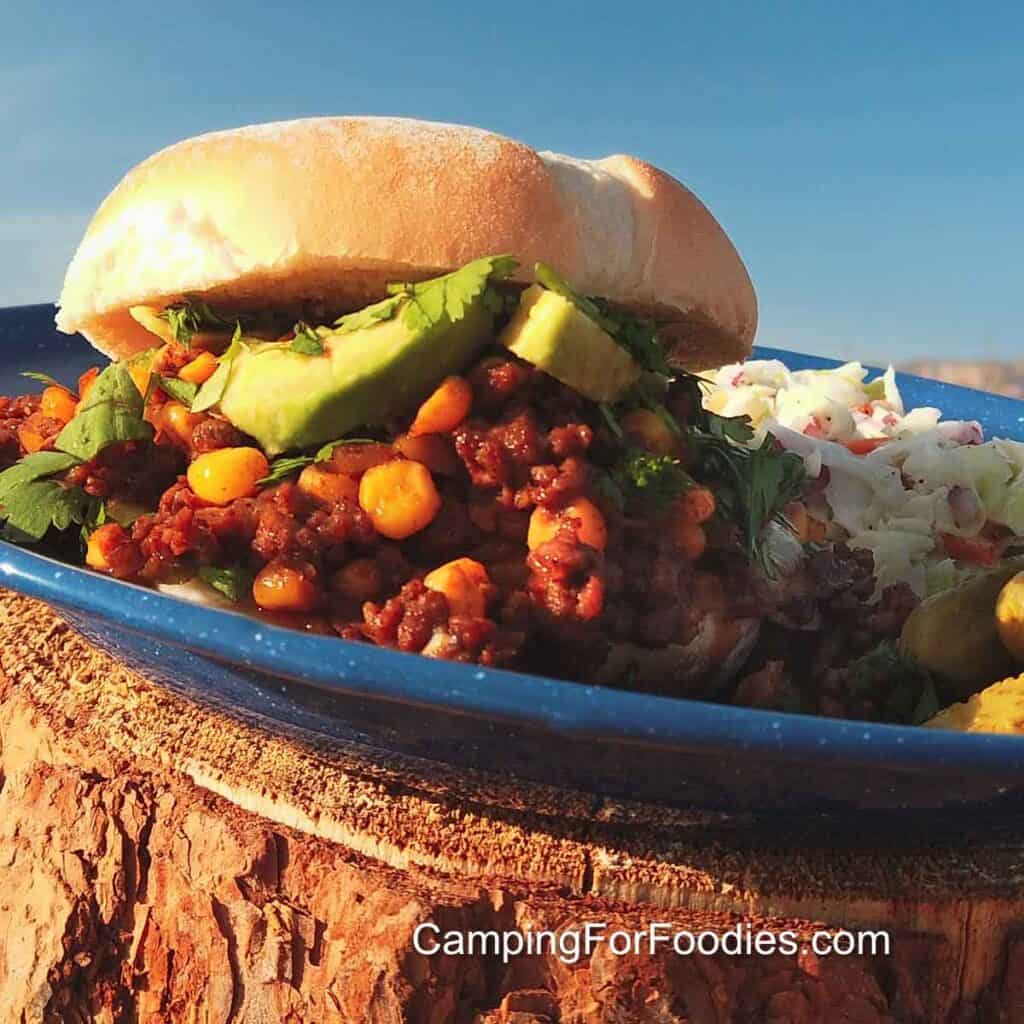 Diablo Sandwich Recipe by CampingForFoodies features a hot sandwich on a blue camping plate. The red and brown color hamburger is mixed with yellow corn, bright green cilantro and avocado slices. The plate is set on a firewood log with blue sky in the background.