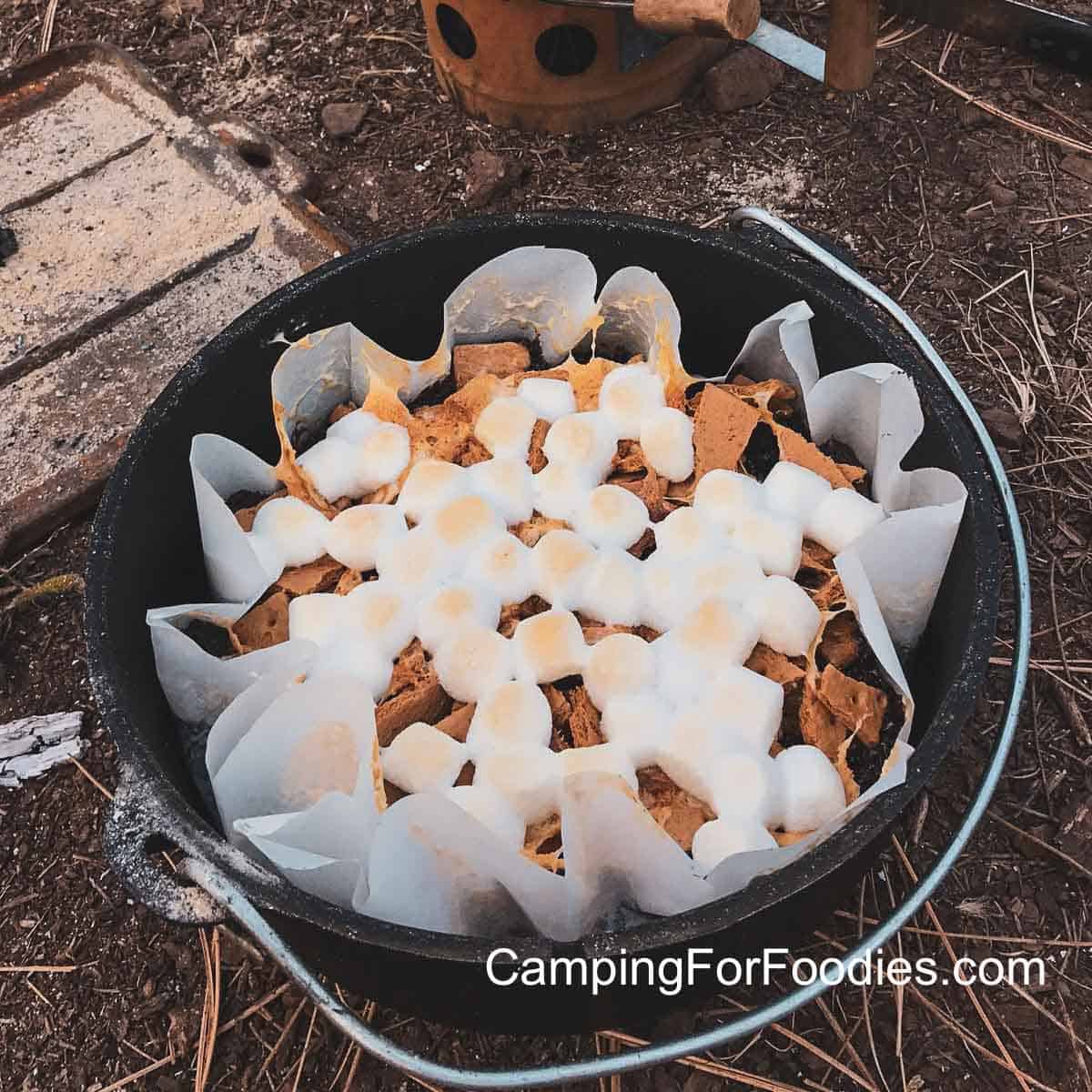 Dutch Oven Brownies by CampingForFoodies features a parchment-paper lined black cast iron camp Dutch oven filled with baked brownies, topped with golden-brown puffed mini marshmallows and graham cracker pieces.